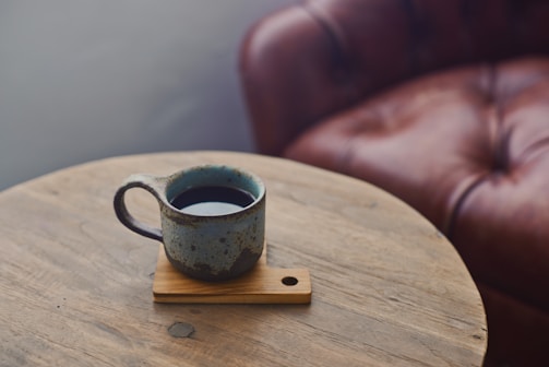 A rustic ceramic mug filled with coffee sits on a wooden coaster on top of a round wooden table. In the background, there is a brown leather couch with visible padding and stitching.