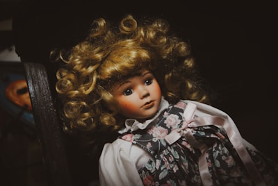 A porcelain doll with curly blonde hair and blue eyes, wearing a floral dress with pink ribbons, is lying on a dark surface. The lighting creates a dramatic contrast, giving the doll an antique appearance.