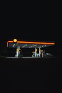 A well-lit gas station stands illuminated against a dark night sky. The bright lights from the station highlight the fueling pumps and create strong contrasts with the surrounding darkness. The Shell logo is visible at the top.