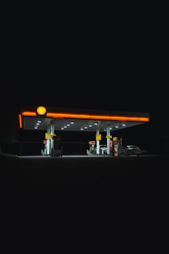A well-lit gas station stands illuminated against a dark night sky. The bright lights from the station highlight the fueling pumps and create strong contrasts with the surrounding darkness. The Shell logo is visible at the top.