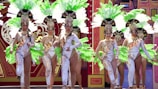 A group of women dressed in elaborate, theatrical costumes perform on stage. Their outfits feature intricate patterns, and they each wear large feathered headdresses in vibrant green and white. The background includes a decorated stage with artistic and colorful designs.
