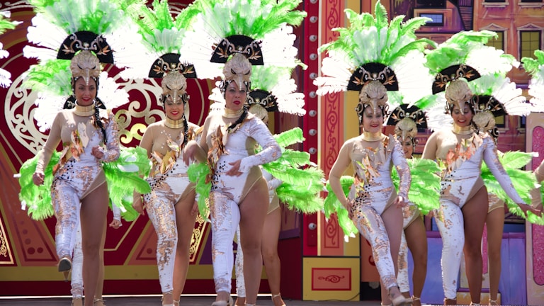A group of women dressed in elaborate, theatrical costumes perform on stage. Their outfits feature intricate patterns, and they each wear large feathered headdresses in vibrant green and white. The background includes a decorated stage with artistic and colorful designs.