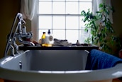 A bathroom scene with a white, classic freestanding bathtub in the foreground. On the edge of the tub rests a blue towel. A bathtub caddy holds various items, including two bottles of bath products, a folded towel, and a decorative object resembling a small duck. In the background, a large window lets in natural light, framed by sheer curtains. To the right of the window is a leafy green plant, adding a touch of nature to the setting.