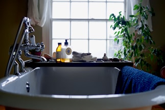 A bathroom scene with a white, classic freestanding bathtub in the foreground. On the edge of the tub rests a blue towel. A bathtub caddy holds various items, including two bottles of bath products, a folded towel, and a decorative object resembling a small duck. In the background, a large window lets in natural light, framed by sheer curtains. To the right of the window is a leafy green plant, adding a touch of nature to the setting.