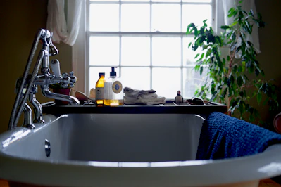 A bathroom scene with a white, classic freestanding bathtub in the foreground. On the edge of the tub rests a blue towel. A bathtub caddy holds various items, including two bottles of bath products, a folded towel, and a decorative object resembling a small duck. In the background, a large window lets in natural light, framed by sheer curtains. To the right of the window is a leafy green plant, adding a touch of nature to the setting.