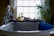 A bathroom scene with a white, classic freestanding bathtub in the foreground. On the edge of the tub rests a blue towel. A bathtub caddy holds various items, including two bottles of bath products, a folded towel, and a decorative object resembling a small duck. In the background, a large window lets in natural light, framed by sheer curtains. To the right of the window is a leafy green plant, adding a touch of nature to the setting.