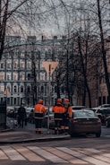 Several construction workers in orange and black uniforms are standing at the edge of a street near a row of parked cars. Leafless trees and a classic multi-story building form the backdrop. A pedestrian with a dog walks on the sidewalk.