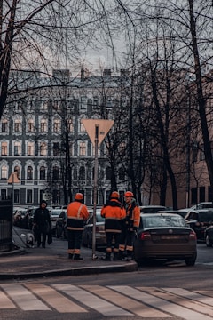 Several construction workers in orange and black uniforms are standing at the edge of a street near a row of parked cars. Leafless trees and a classic multi-story building form the backdrop. A pedestrian with a dog walks on the sidewalk.
