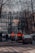 Several construction workers in orange and black uniforms are standing at the edge of a street near a row of parked cars. Leafless trees and a classic multi-story building form the backdrop. A pedestrian with a dog walks on the sidewalk.