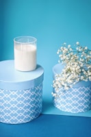 A decorative arrangement features two cylindrical blue gift boxes with a white geometric pattern on the sides. A glass candle holder with a white candle is placed on top of one box. A small bunch of delicate white flowers, possibly baby's breath, drape over the edge of the other box. Both boxes and the items sit against a matching blue background.