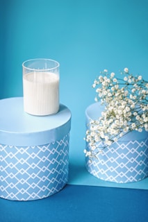 A decorative arrangement features two cylindrical blue gift boxes with a white geometric pattern on the sides. A glass candle holder with a white candle is placed on top of one box. A small bunch of delicate white flowers, possibly baby's breath, drape over the edge of the other box. Both boxes and the items sit against a matching blue background.