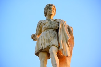 A stone statue of a figure dressed in Renaissance-style clothing stands prominently against a clear blue sky. The figure has curly hair and holds a draped garment in one hand, exuding a sense of movement and grace.
