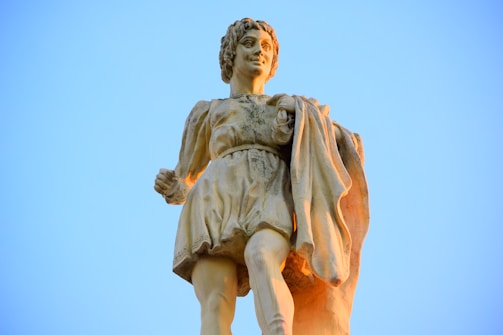A stone statue of a figure dressed in Renaissance-style clothing stands prominently against a clear blue sky. The figure has curly hair and holds a draped garment in one hand, exuding a sense of movement and grace.