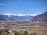 A vast agricultural landscape stretches towards a range of snow-capped mountains under a clear blue sky. The fields are meticulously organized, possibly vineyards or orchards, surrounded by small buildings and patches of greenery.