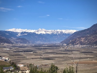 A vast agricultural landscape stretches towards a range of snow-capped mountains under a clear blue sky. The fields are meticulously organized, possibly vineyards or orchards, surrounded by small buildings and patches of greenery.