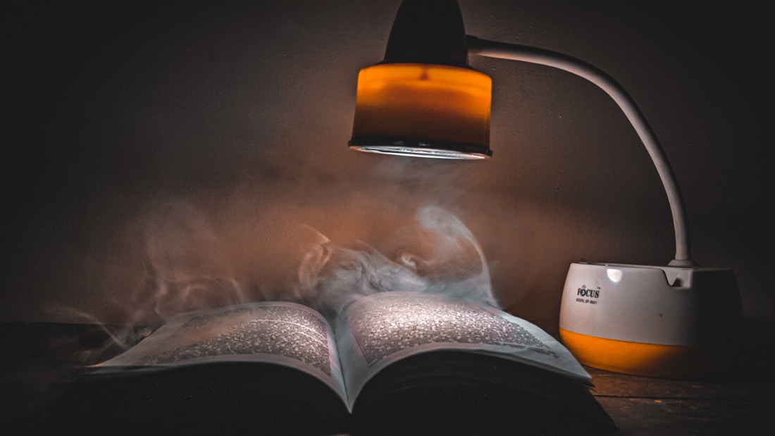 An open book lies on a surface with visible light shining on it from a small lamp. Wisps of smoke or mist swirl around the book, creating a mysterious atmosphere. The lamp has a modern design with a curved neck and an orange accent, providing a warm glow that contrasts with the dark, moody background.