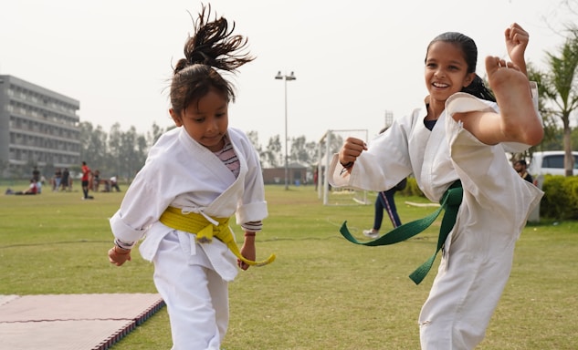 Two children wearing martial arts uniforms practice in a grassy outdoor area. One child, with a yellow belt, appears to be in a defensive stance, while the other, with a green belt, executes a high kick. In the background, a multi-story building and some trees are visible, with additional people in the distance.