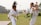 Two children wearing martial arts uniforms practice in a grassy outdoor area. One child, with a yellow belt, appears to be in a defensive stance, while the other, with a green belt, executes a high kick. In the background, a multi-story building and some trees are visible, with additional people in the distance.