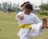 A child wearing a white martial arts uniform with a yellow belt performs a high kick in a grassy outdoor area. The child's facial expression shows concentration and effort. In the background, there are trees and a few people, suggesting a park or open field setting.