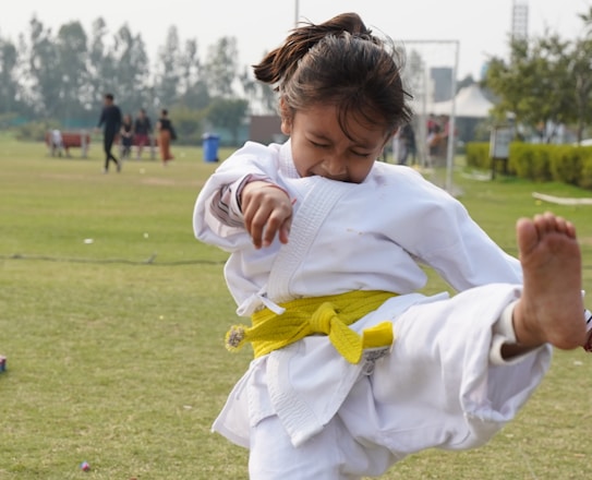 A child wearing a white martial arts uniform with a yellow belt performs a high kick in a grassy outdoor area. The child's facial expression shows concentration and effort. In the background, there are trees and a few people, suggesting a park or open field setting.