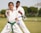 Two individuals are practicing martial arts outdoors, wearing white uniforms with colored belts. The younger person in the foreground has a green belt and is executing a punch with determination, while the older person in the background has a blue belt and observes attentively. The scene takes place on a grassy field with trees and a partly cloudy sky.