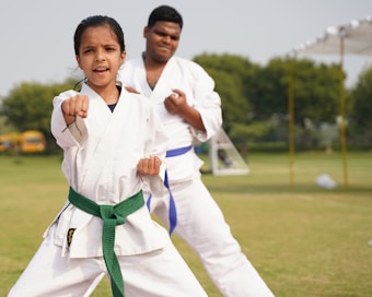 Two individuals are practicing martial arts outdoors, wearing white uniforms with colored belts. The younger person in the foreground has a green belt and is executing a punch with determination, while the older person in the background has a blue belt and observes attentively. The scene takes place on a grassy field with trees and a partly cloudy sky.