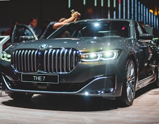 A sleek, high-end grey sedan with a distinctive grille and illuminated headlights is prominently displayed. Reflections and light flares enhance the car's polished appearance. A few people are visible in the background near the open driver's side door.