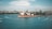 Aerial view of the Sydney Opera House situated on the waterfront, surrounded by several boats sailing in the harbor. The skyline of Sydney with numerous buildings forms the backdrop, under a partly cloudy sky.