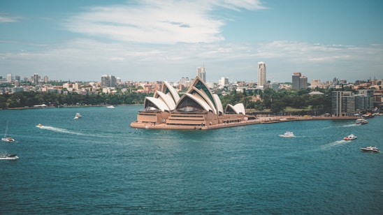 Aerial view of the Sydney Opera House situated on the waterfront, surrounded by several boats sailing in the harbor. The skyline of Sydney with numerous buildings forms the backdrop, under a partly cloudy sky.