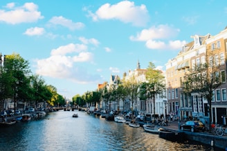 A scenic canal lined with historic buildings and lush green trees under a bright blue sky with fluffy white clouds. Several boats are docked along the waterway, and people can be seen walking or cycling alongside the picturesque canal.