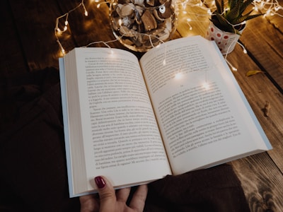 An open book with text in Italian is resting on a wooden table. Soft, warm fairy lights are draped around, casting a cozy glow. A clear jar filled with wood chips and a small potted plant are nearby, adding to the rustic ambiance.