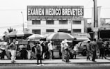 A group of people stand on a sidewalk beside several street vendors with umbrellas. Above them, a large sign advertises medical exams and license testing. Buildings and various advertisements are visible in the background.