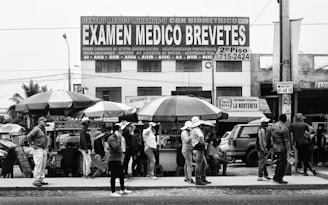 A group of people stand on a sidewalk beside several street vendors with umbrellas. Above them, a large sign advertises medical exams and license testing. Buildings and various advertisements are visible in the background.