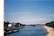 A serene river flows through an urban area with a sandy beach lining one side. Several people enjoy the water and the beach. Buildings and greenery are visible along the riverbank under a clear blue sky. A strip of light leaks on the left side of the image, likely from a camera film defect.