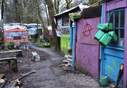 A series of colorful, rustic structures and caravans line a narrow, winding path through a wooded area. The buildings display vibrant hues of purple, blue, and green, with noticeable signs of wear and graffiti, including red anarchist symbols. Green fuel cans hang from a door, and various objects such as plants, benches, and containers are scattered around. The setting feels makeshift and artistic.