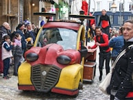 A whimsical, colorful vehicle resembling a cartoon character drives through a busy street. Performers in red and black outfits interact energetically with onlookers, creating a lively and festive atmosphere. The crowd, including families with children, appears engaged and entertained.