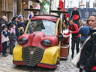 A whimsical, colorful vehicle resembling a cartoon character drives through a busy street. Performers in red and black outfits interact energetically with onlookers, creating a lively and festive atmosphere. The crowd, including families with children, appears engaged and entertained.