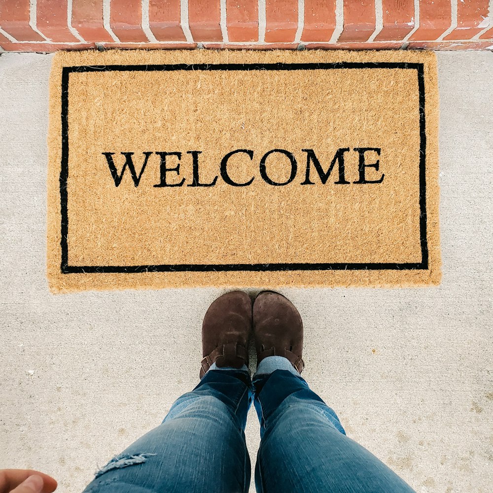 Persona en jeans vaqueros azules de pie sobre una alfombra de bienvenida marrón y negra