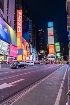 A bustling urban scene featuring bright neon lights and towering digital billboards. The street is lined with advertisements for various brands, and there are several vehicles, including taxis, driving along the road. Pedestrians can be seen walking in the background, adding to the vibrant atmosphere.