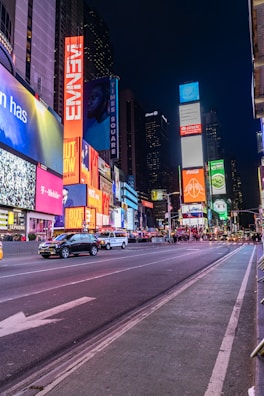 A bustling urban scene featuring bright neon lights and towering digital billboards. The street is lined with advertisements for various brands, and there are several vehicles, including taxis, driving along the road. Pedestrians can be seen walking in the background, adding to the vibrant atmosphere.