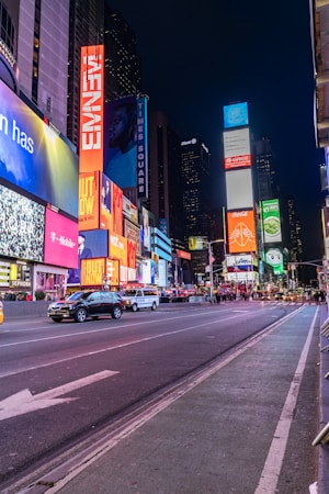 A bustling urban scene featuring bright neon lights and towering digital billboards. The street is lined with advertisements for various brands, and there are several vehicles, including taxis, driving along the road. Pedestrians can be seen walking in the background, adding to the vibrant atmosphere.