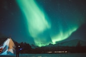 A mesmerizing display of the Northern Lights illuminates the night sky above snow-covered mountains and a traditional tent. The vibrant green and turquoise hues of the aurora contrast with the dark, starry sky, creating a breathtaking natural spectacle.