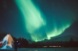 A mesmerizing display of the Northern Lights illuminates the night sky above snow-covered mountains and a traditional tent. The vibrant green and turquoise hues of the aurora contrast with the dark, starry sky, creating a breathtaking natural spectacle.