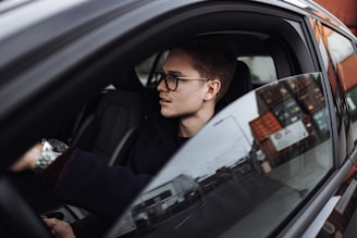 A person wearing glasses is seated inside a car, appearing to focus intently ahead. The car window is partially down, revealing reflections of an urban environment with buildings and signs outside.