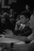 Several people appear to be attentively participating in a discussion or meeting, seated in a dimly lit environment with focus on one individual deep in thought.