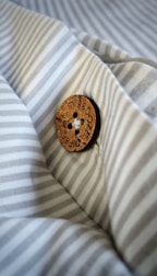 A wooden button with the inscription 'DIP & DOZE' is sewn onto a piece of fabric with thin, light grey and white stripes. The button is detailed and has four holes sewn with white thread, creating a contrast against the fabric's pattern.