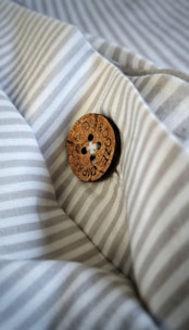 A wooden button with the inscription 'DIP & DOZE' is sewn onto a piece of fabric with thin, light grey and white stripes. The button is detailed and has four holes sewn with white thread, creating a contrast against the fabric's pattern.