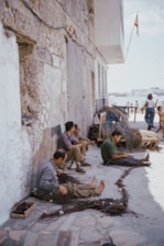Several men are sitting on the ground against an old stone wall, working on fishing nets. In the background, a couple of women are walking near the beach with a visible building structure. There is a pile of fishing nets and some wooden boxes nearby. A flag is mounted on a pole extending from the building.