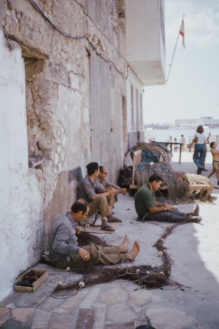 Several men are sitting on the ground against an old stone wall, working on fishing nets. In the background, a couple of women are walking near the beach with a visible building structure. There is a pile of fishing nets and some wooden boxes nearby. A flag is mounted on a pole extending from the building.
