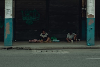 Two people sit on a sidewalk under a sheltered area in front of closed metal shutters. They appear to be surrounded by bags and blankets, suggesting they might be experiencing homelessness. The overall atmosphere is quiet and somber.